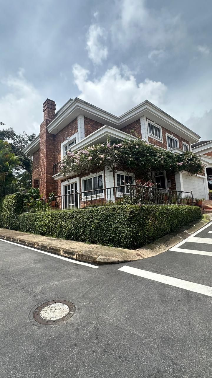 Paisaje urbano con edificios y árboles verdes, montañas al fondo bajo un cielo nublado.