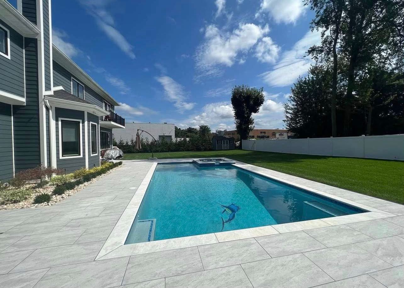 A rectangular pool with turquoise water, surrounded by gray paving, in a backyard next to a dark gray house.