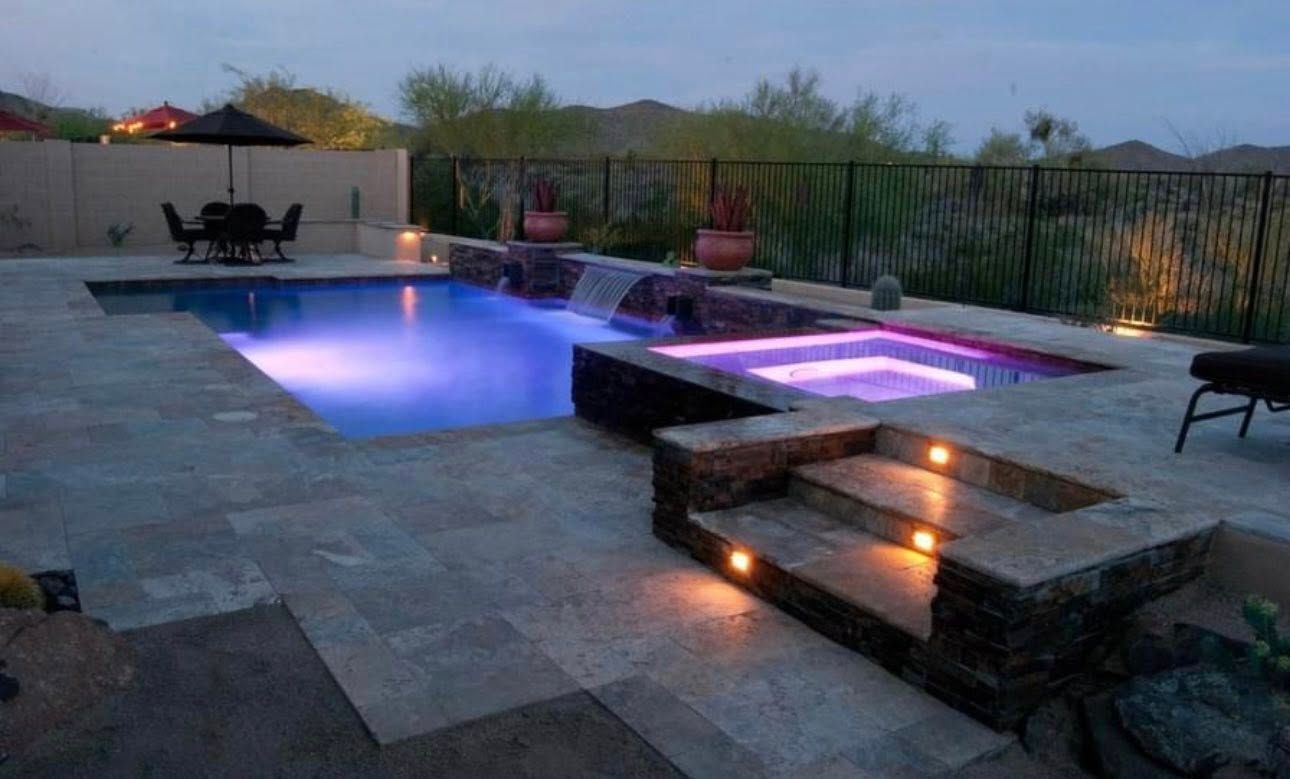Night view of a pool with a lit jacuzzi, steps, and patio in a backyard setting.