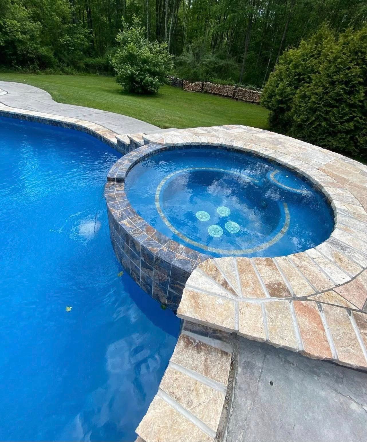Blue swimming pool and attached hot tub with stone and brick surround. Lush green lawn in background.
