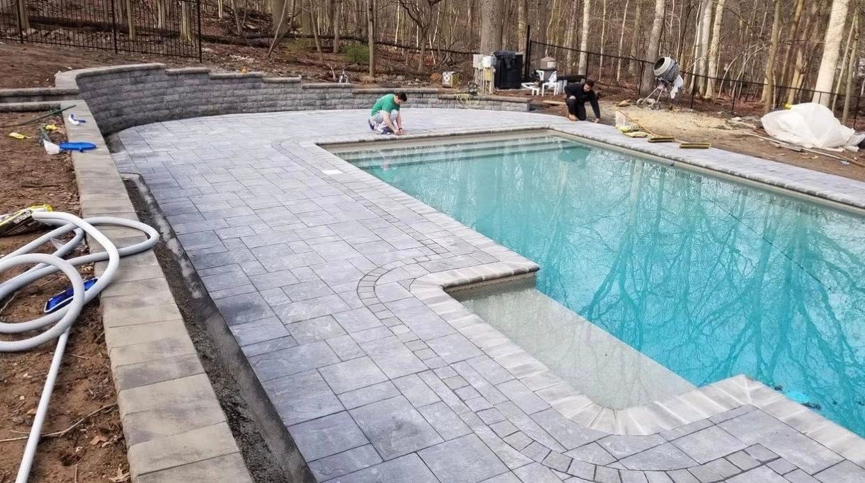 Pool with gray pavers and steps under construction; people nearby in a wooded setting.