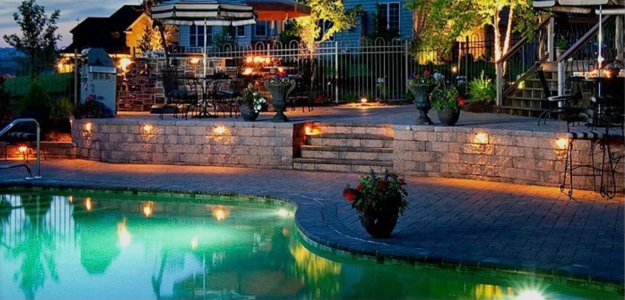 Night view of an illuminated pool, patio, and raised stone wall; house and trees in background.