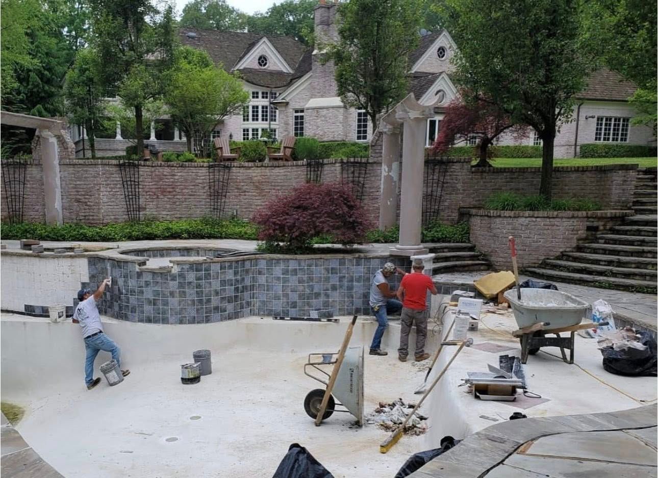 Pool under construction; workers installing tiles. White pool, brick wall, large house in background.