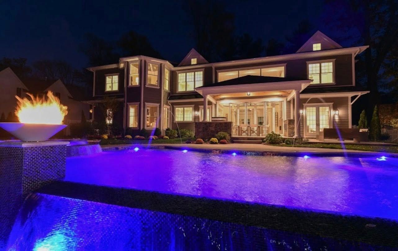Night view of a luxury home with a lit pool, fire pit, and blue lights reflecting on the water.