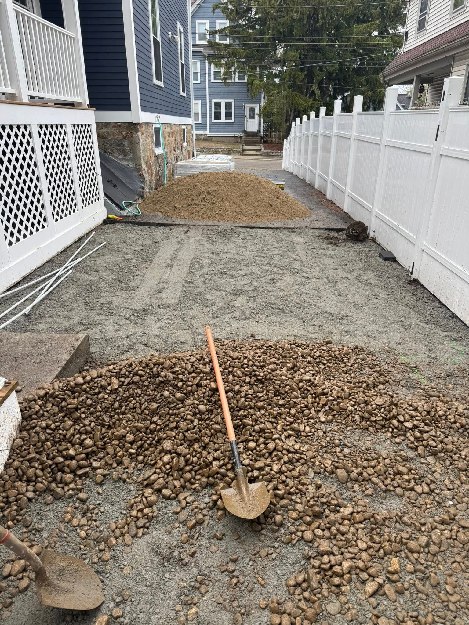 A gravel and sand driveway under construction, with a shovel in the center, flanked by white fences and homes.