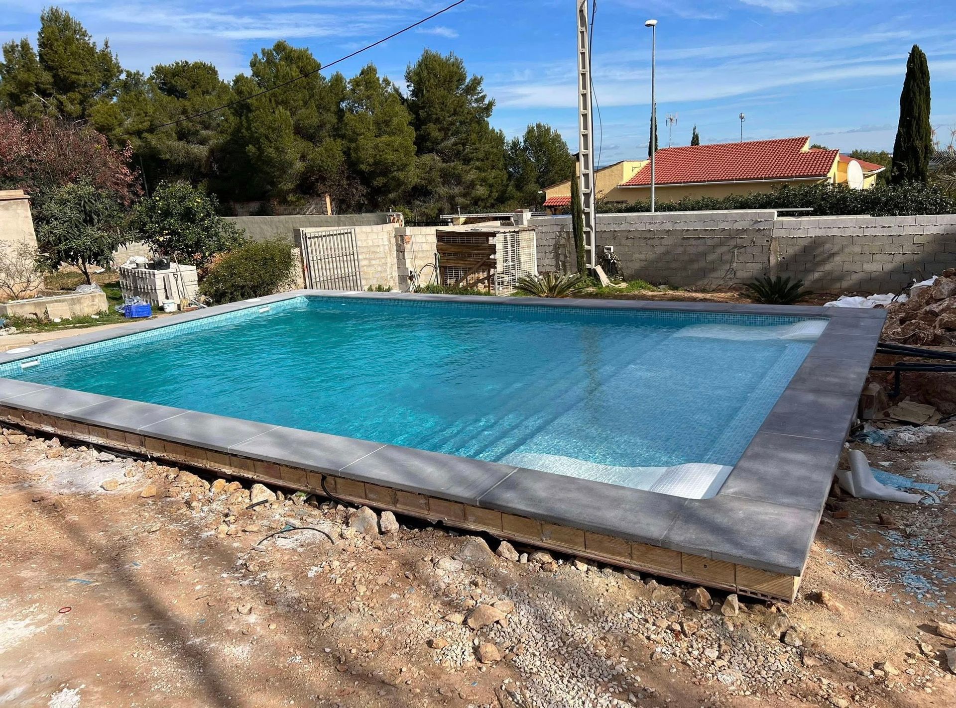 Rectangular swimming pool with blue water, concrete edging, and surrounding dirt ground.