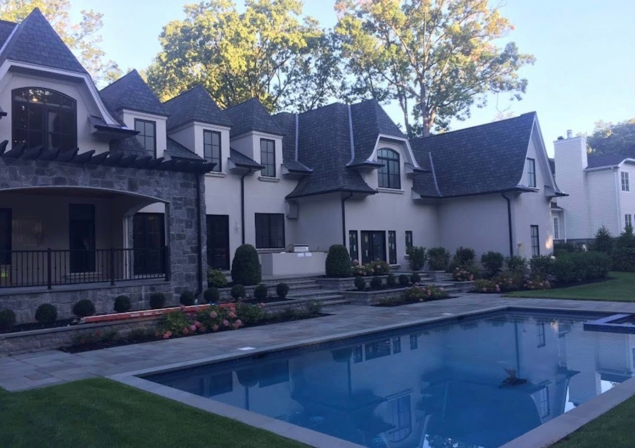 Large house with pool, stone and stucco exterior, dark roof, and lush landscaping.