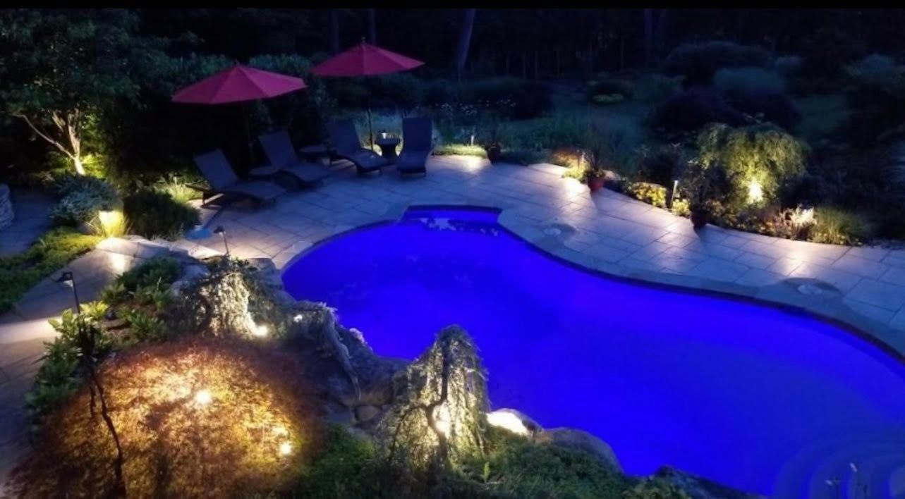 Nighttime view of a pool with blue water and surrounding landscape lighting. Lounge chairs and red umbrellas are visible.