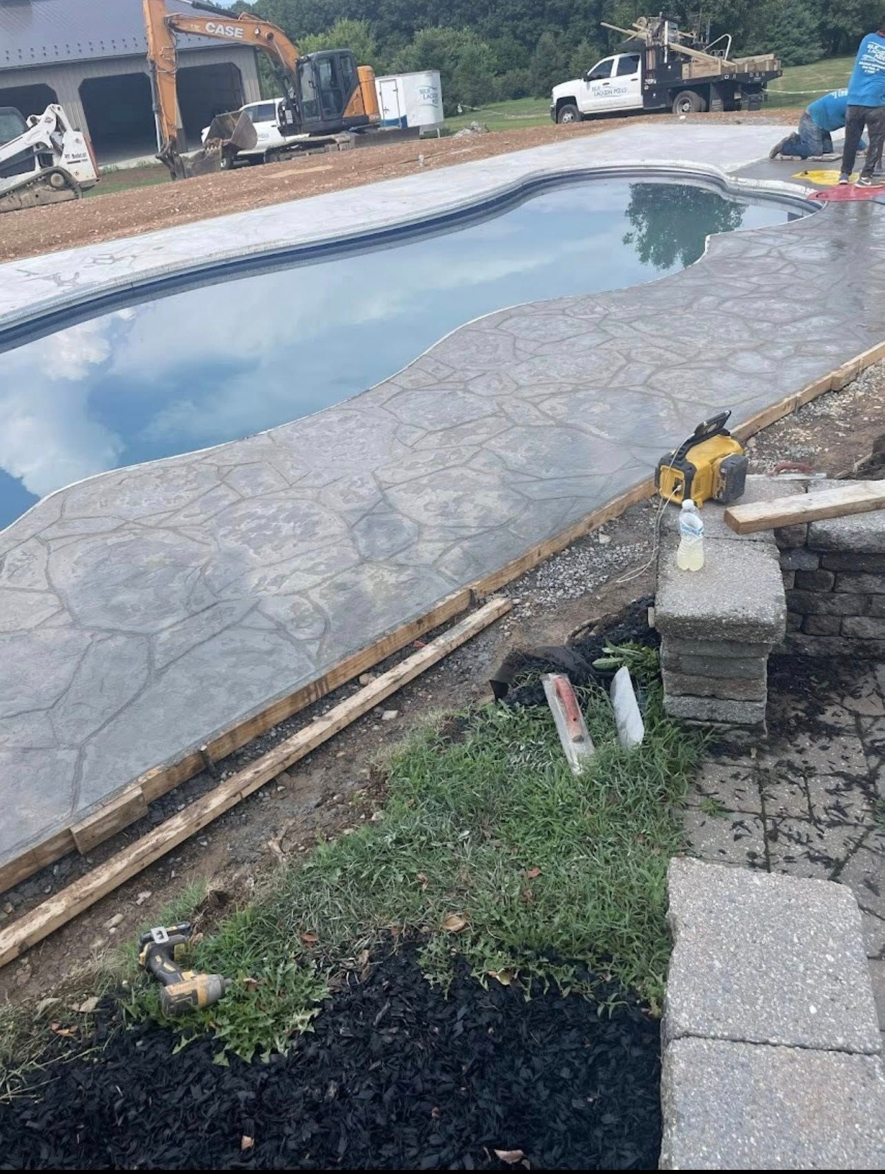 Poolside construction: concrete deck, pool, landscaping, and construction equipment in the background.