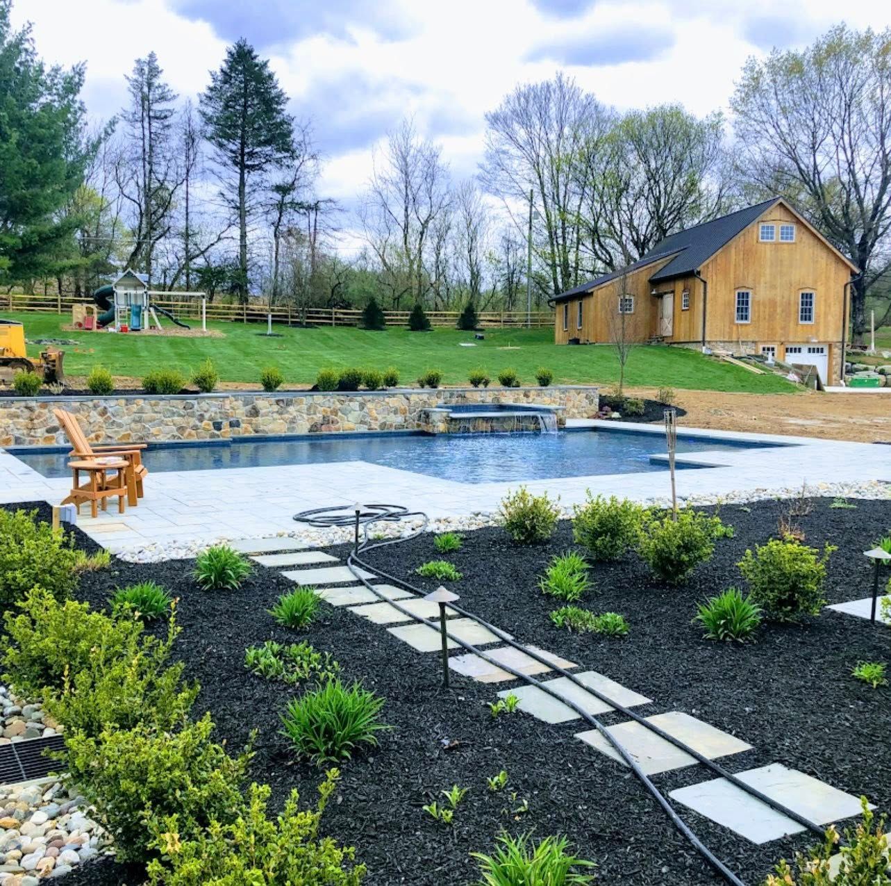 Backyard with pool, stone patio, landscaping, and wood-sided house under cloudy sky.