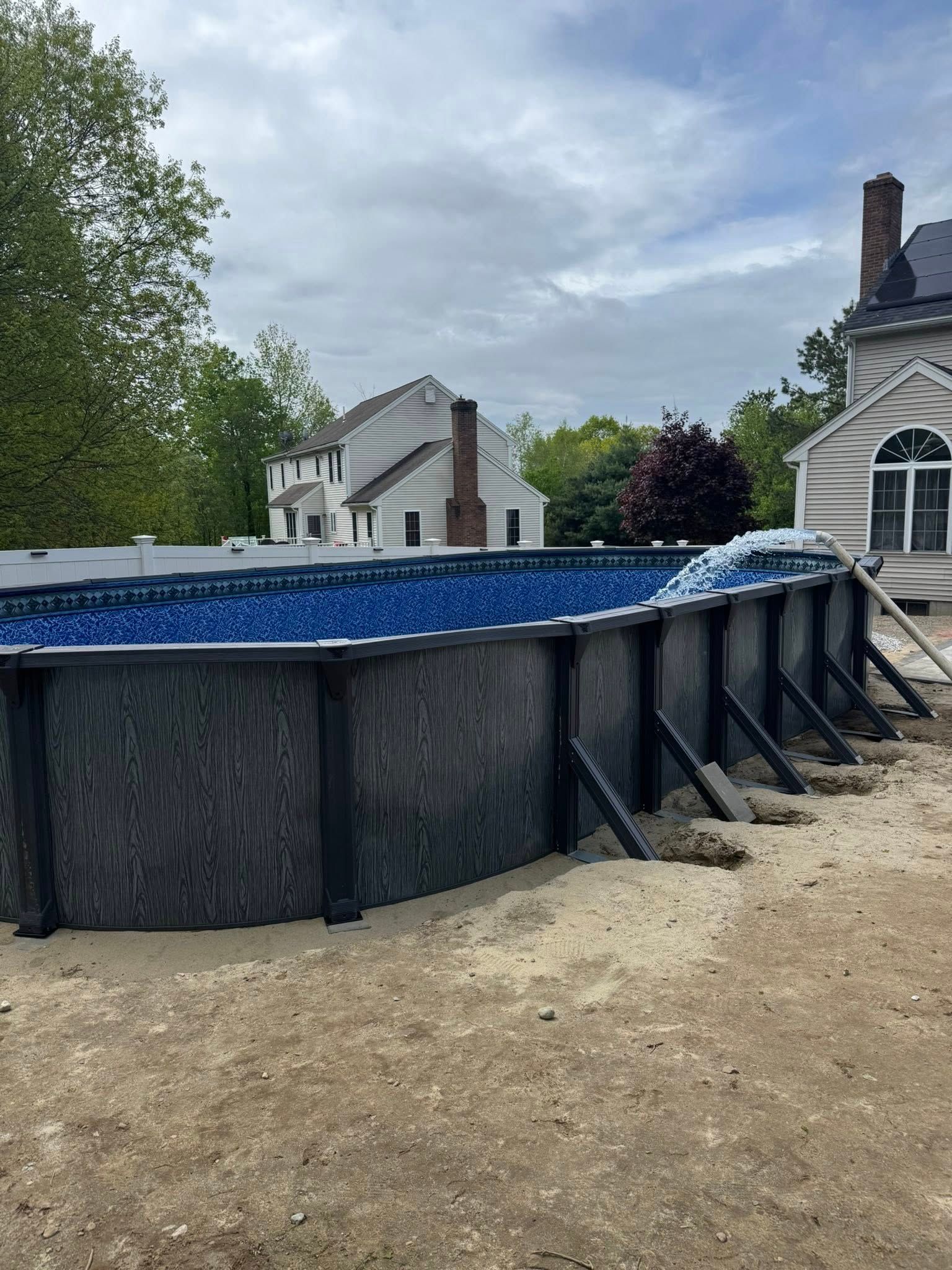 Above-ground swimming pool under construction. Wooden frame with blue interior. Sandy ground, cloudy sky.
