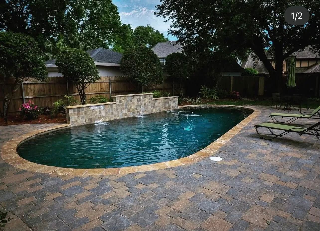 Oval swimming pool with stone waterfall, surrounded by brick patio, trees and lawn chairs.