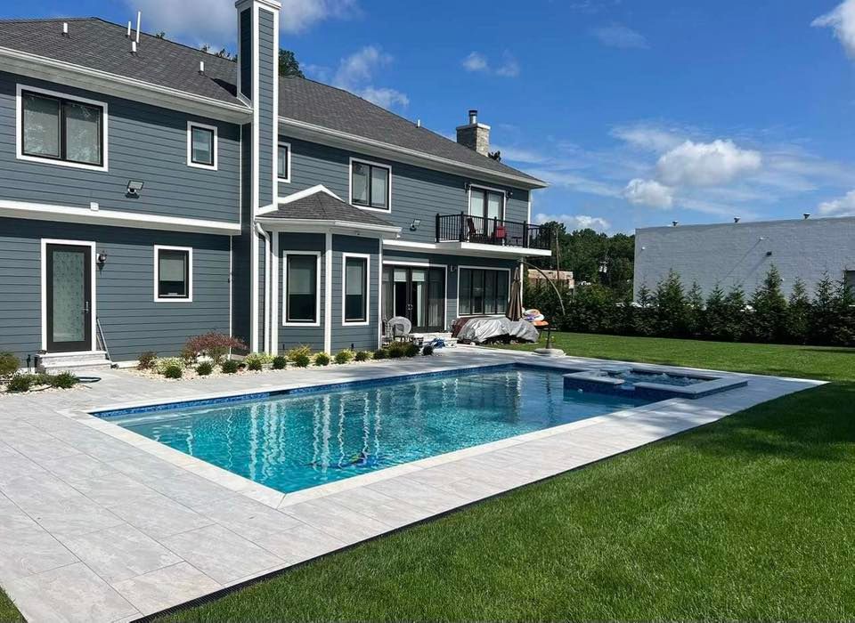 Backyard with pool, spa, and two-story house with blue siding. White stone patio and green lawn.