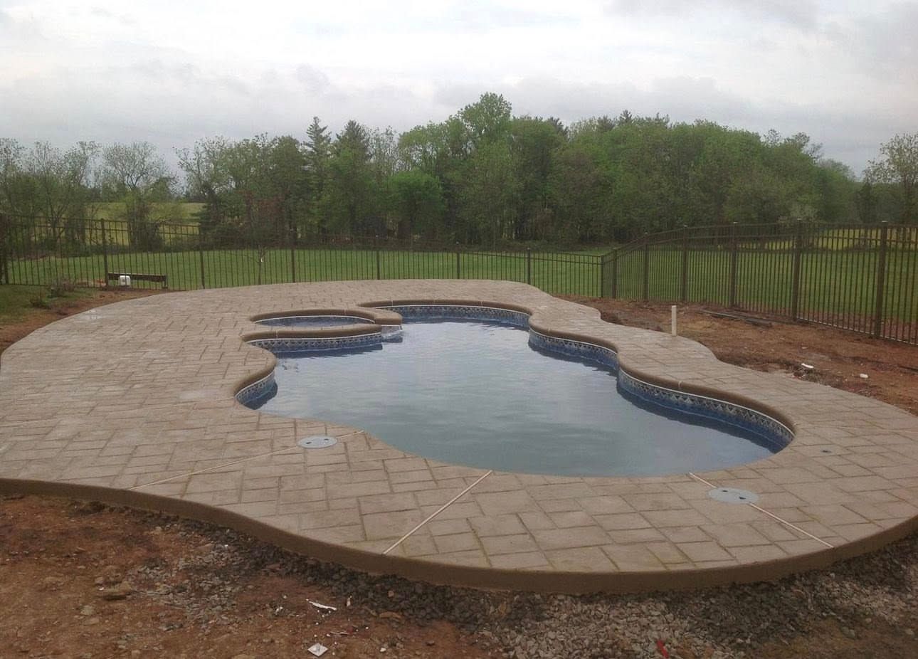 Pool with stone patio and wooden fence in a rural setting, overcast sky.