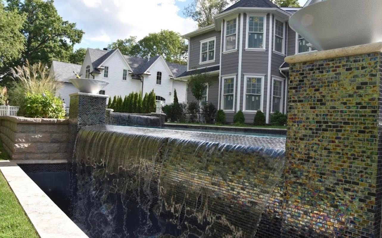 Water cascades over a pool edge into a dark basin; gray house with turret in background.