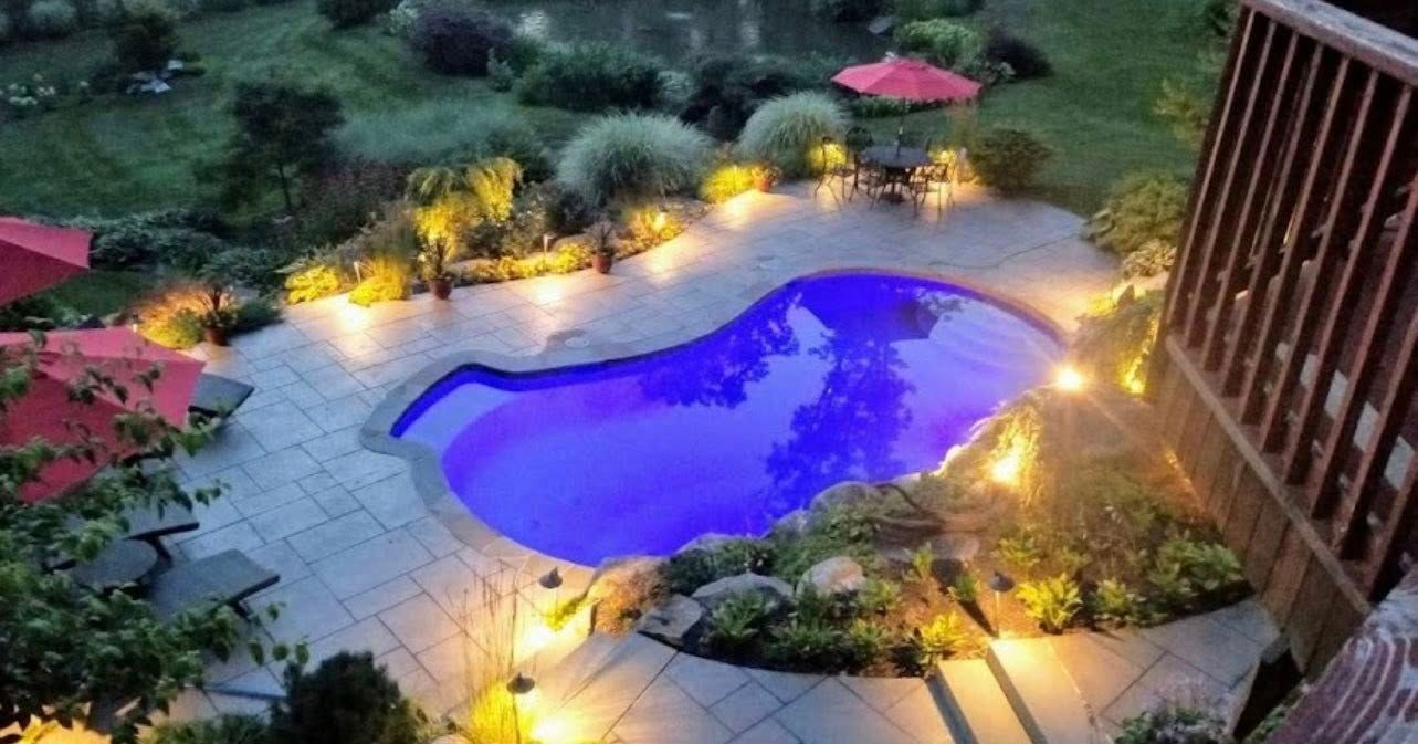 Night view of a kidney-shaped pool illuminated by lights, with outdoor seating and lush landscaping.