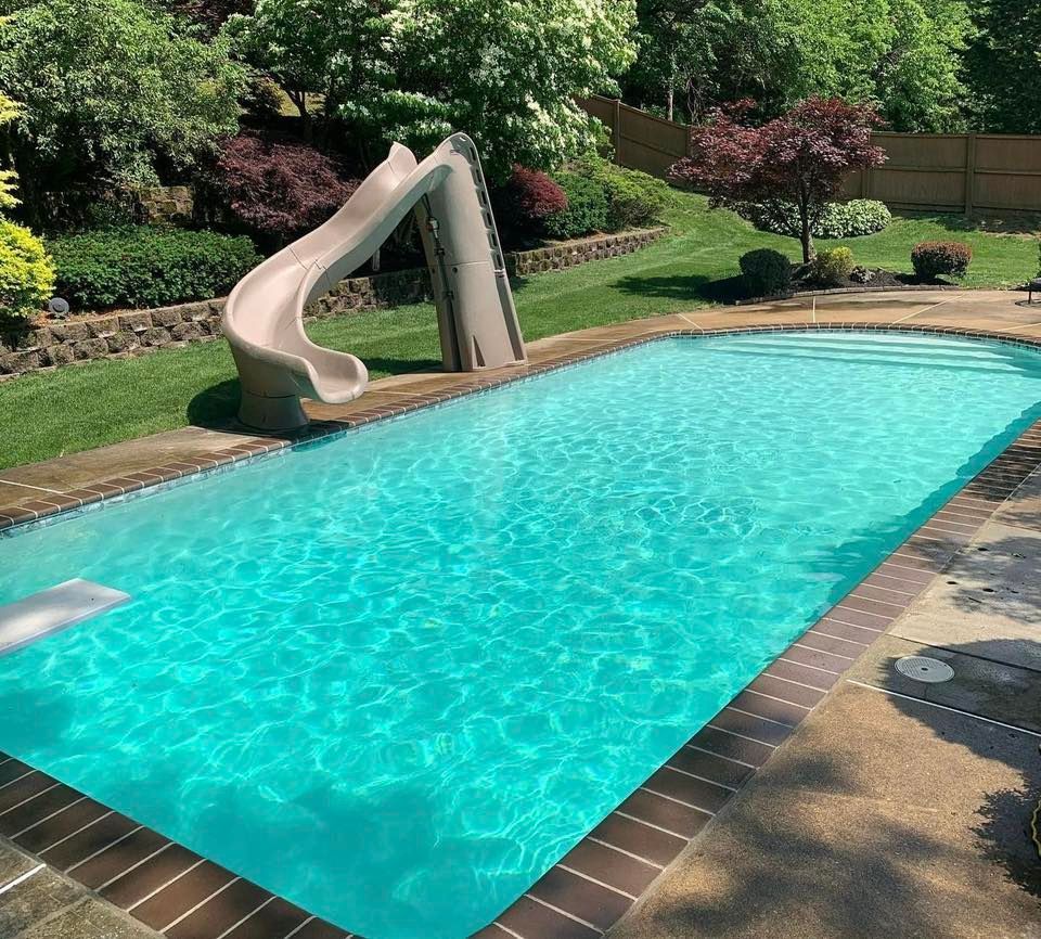 Rectangular pool with slide in a well-manicured backyard with lush greenery and a brick border.