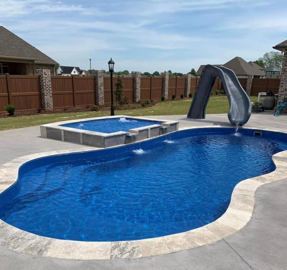 Blue pool with attached spa, slide, and concrete patio. Brown fence and house in background.