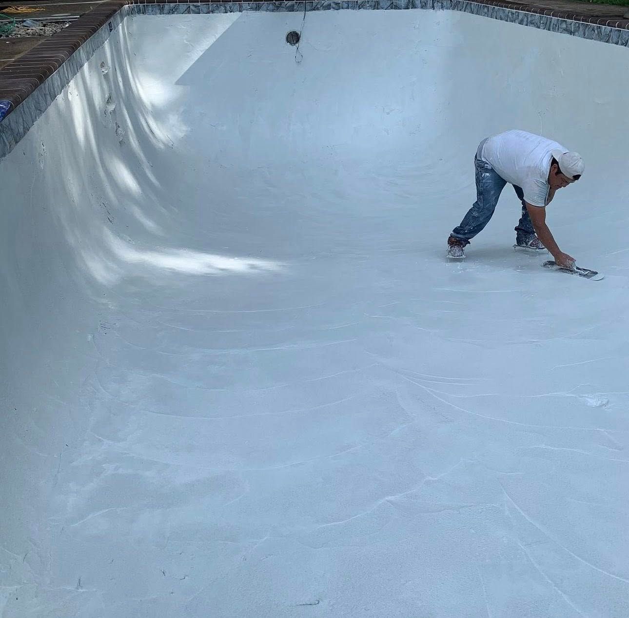 Person applying white sealant to the interior of an empty swimming pool.