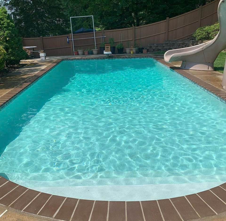 Rectangular pool filled with turquoise water, brown brick border. Slide on right, trees and fence in background.