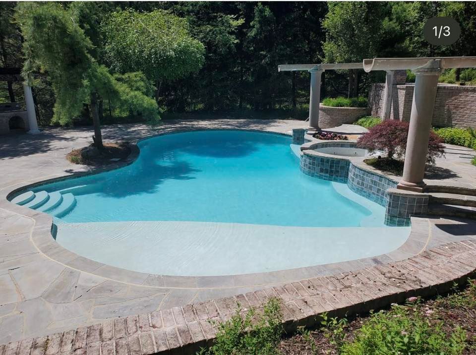 Swimming pool with light blue water and stone patio surrounded by trees.