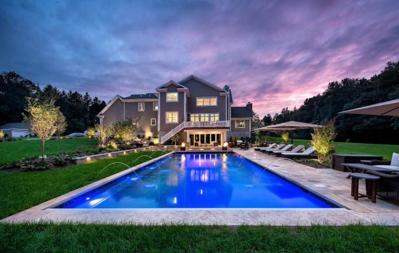 Luxury home with a rectangular pool illuminated blue at dusk; loungers, outdoor seating.