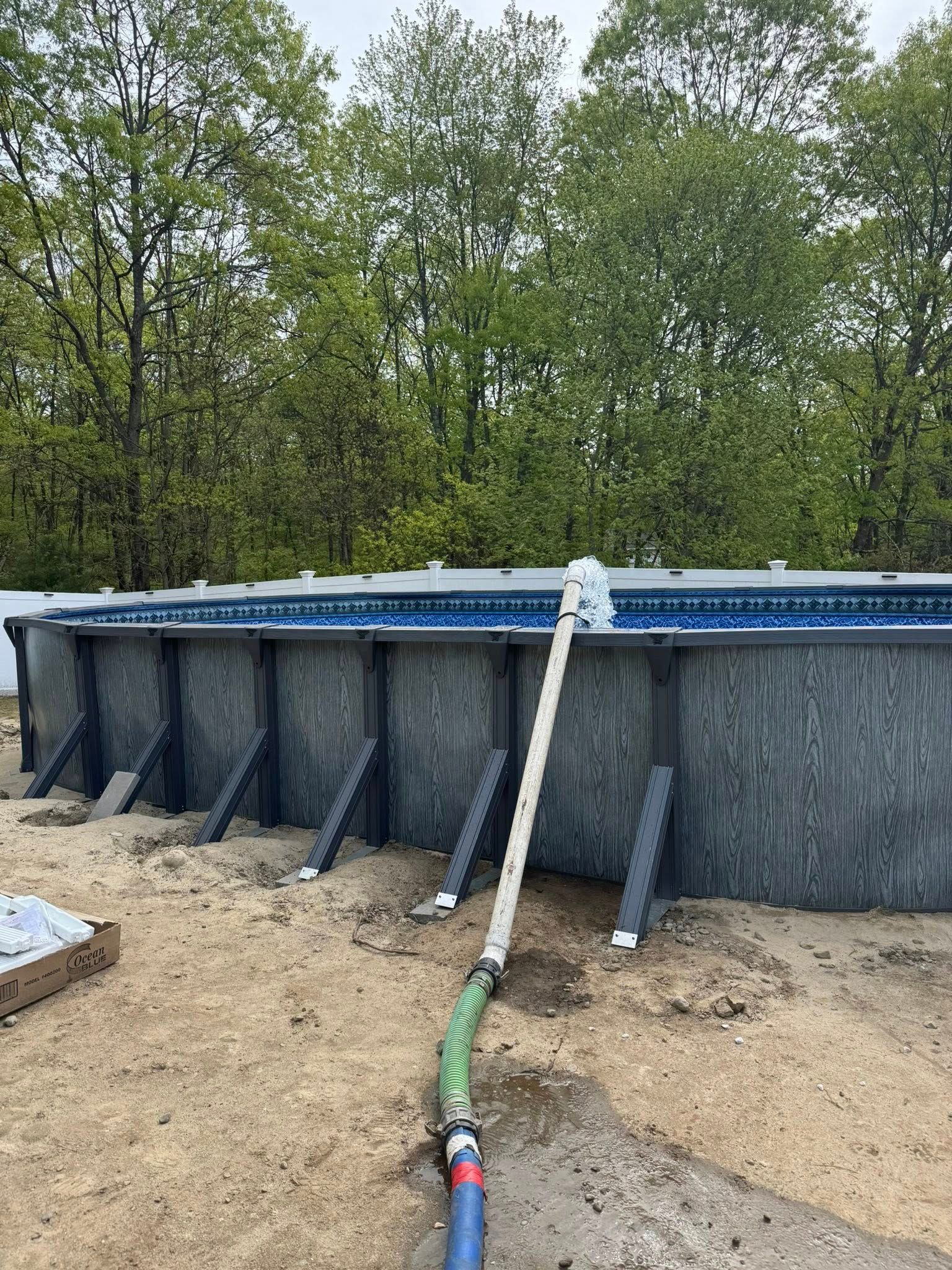 Above-ground pool under construction, with plumbing and support beams visible.  Trees and overcast sky in the background.