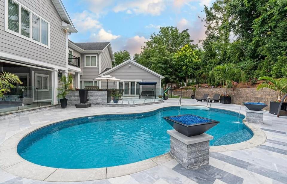 Backyard with a swimming pool, stone patio, and two-story house. Blue water, blue fire pit. Sunny sky.