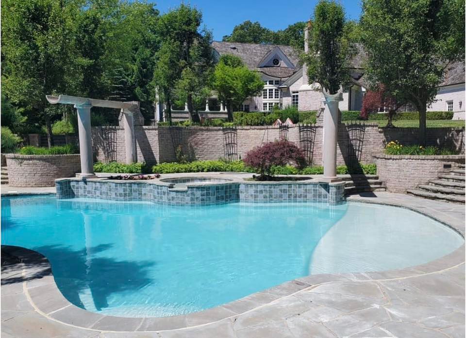 Blue swimming pool with stone deck, pillars, and a multi-story house in the background.