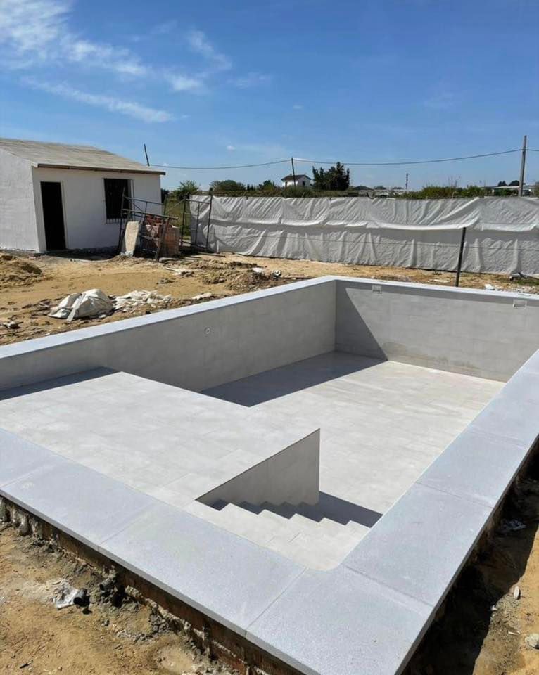 Construction site with a rectangular pool under construction, house in background, clear sky.
