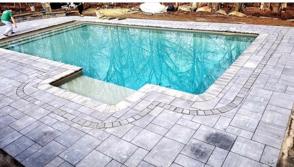 Pool with light blue water and stone patio being installed; person walks nearby.