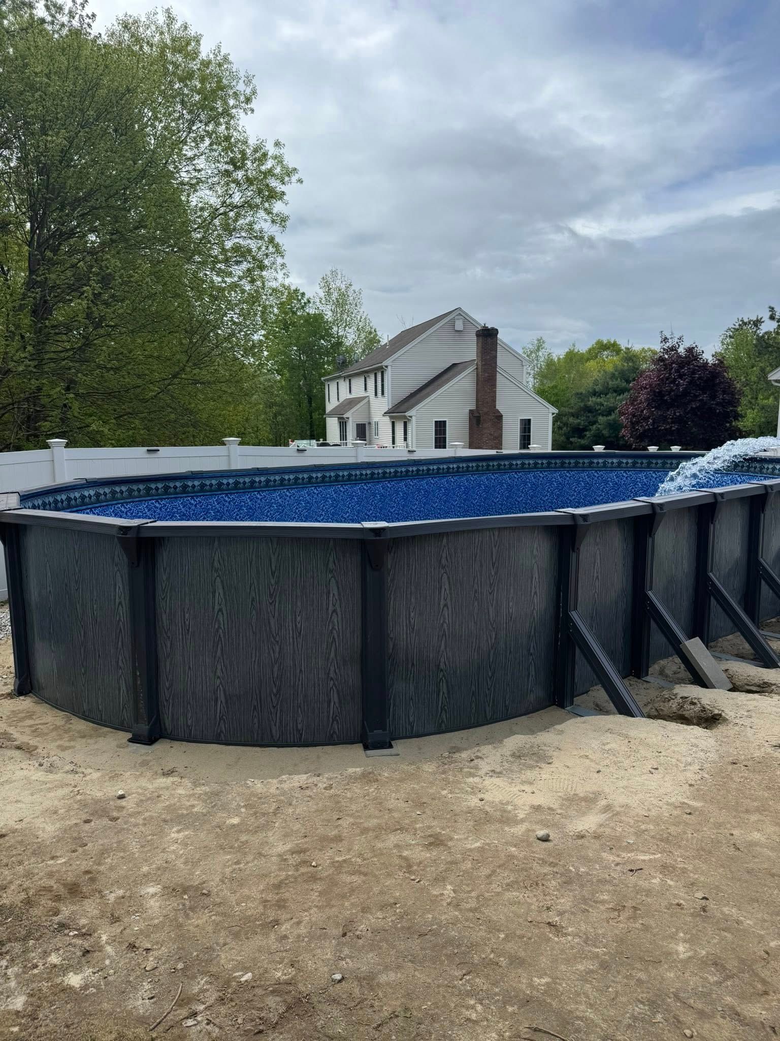 Above-ground pool construction: A dark-paneled pool surrounded by sand, with a house and cloudy sky in the background.