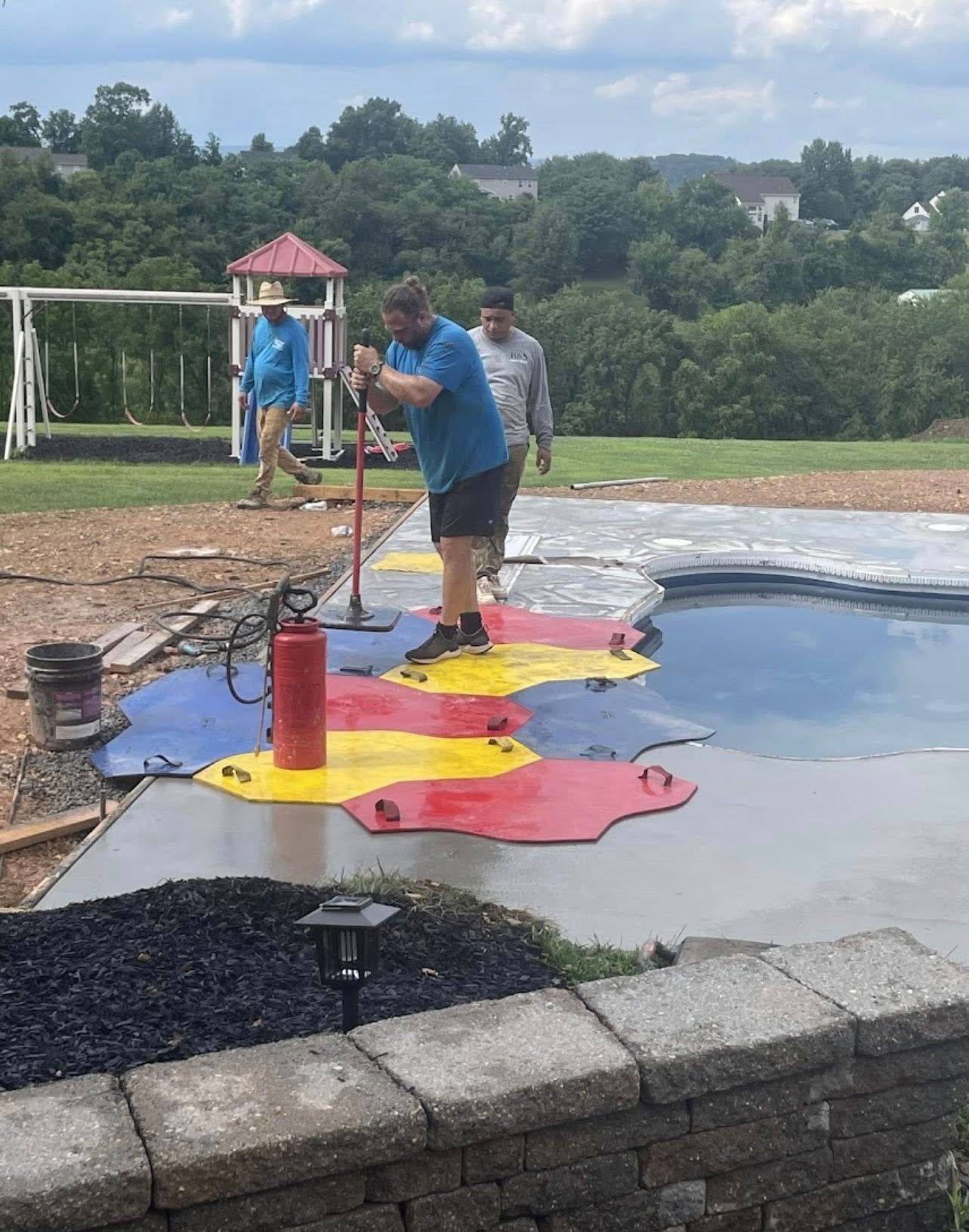 Three workers laying colorful tiles around a pool. A man leans on a tool. Outdoors, daytime.