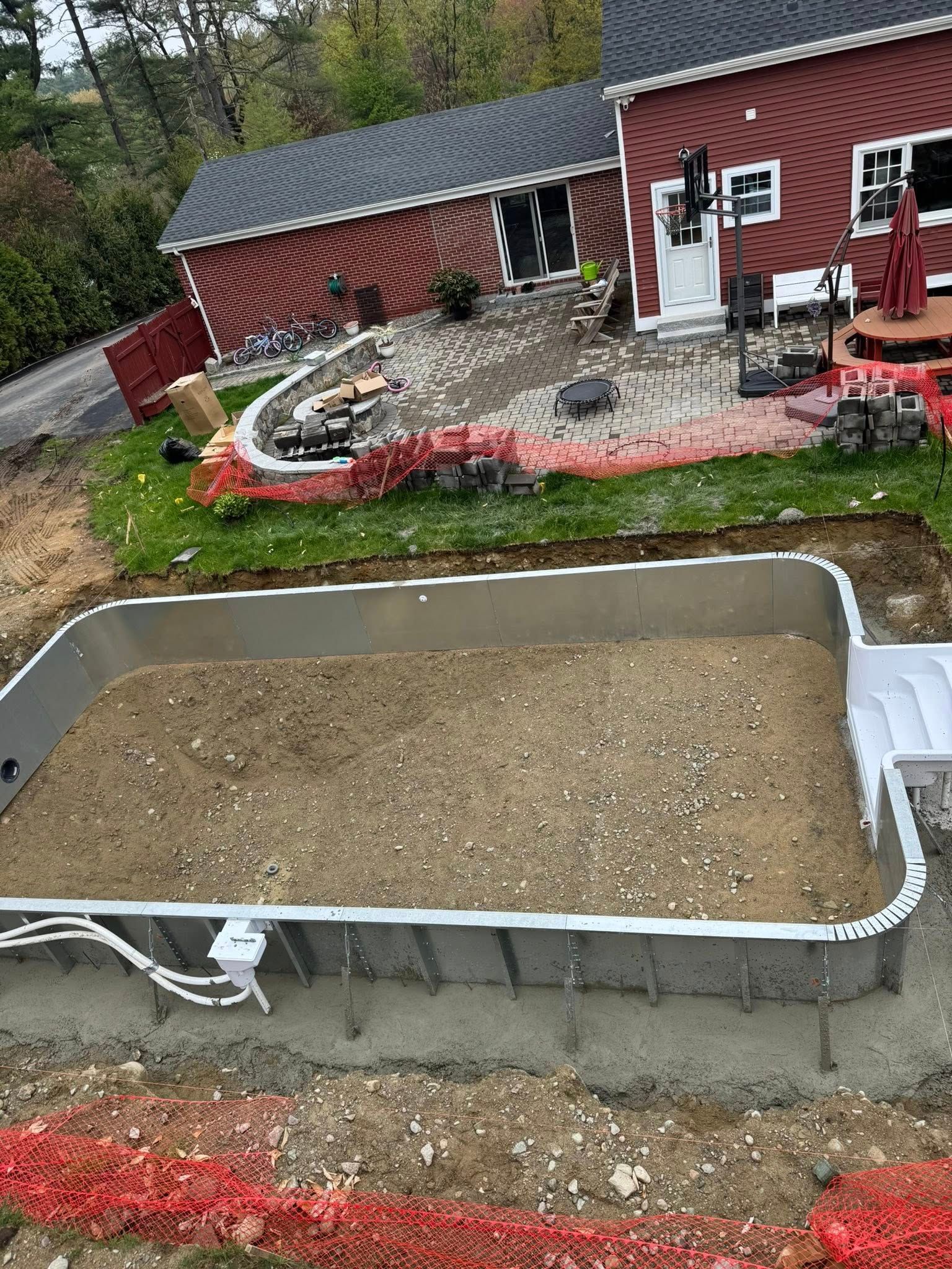 An in-ground pool under construction in a yard. Gravel fill inside the pool, red house and patio in background.
