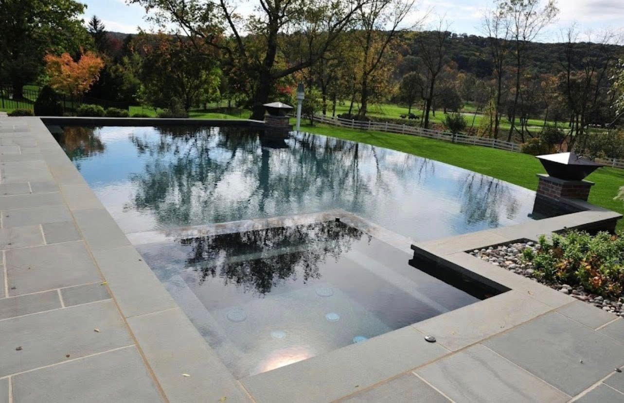 Rectangular swimming pool reflecting trees and sky, surrounded by stone patio and lawn.