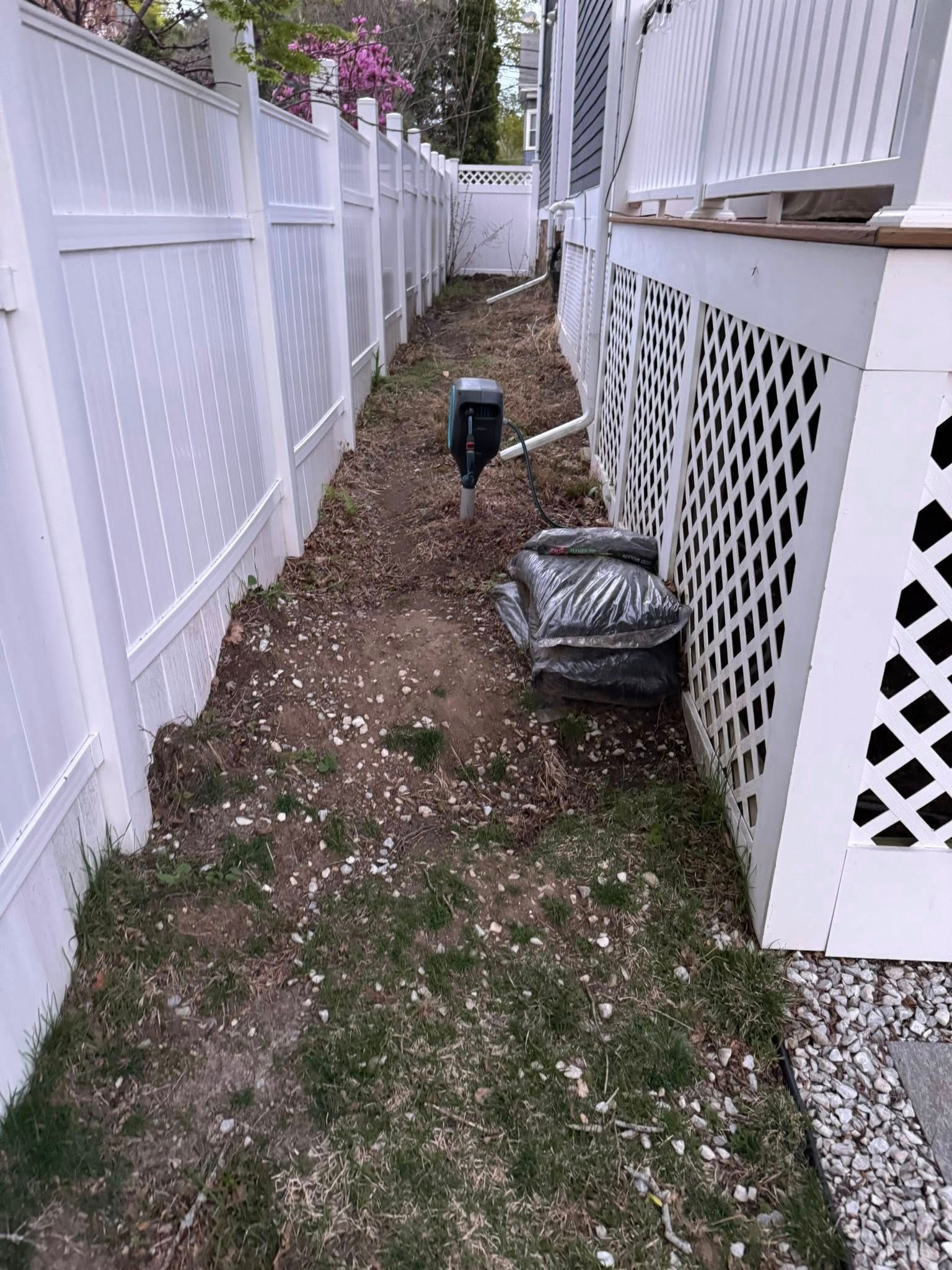 Narrow, dirt and grass yard between white fences and house with bags of mulch.