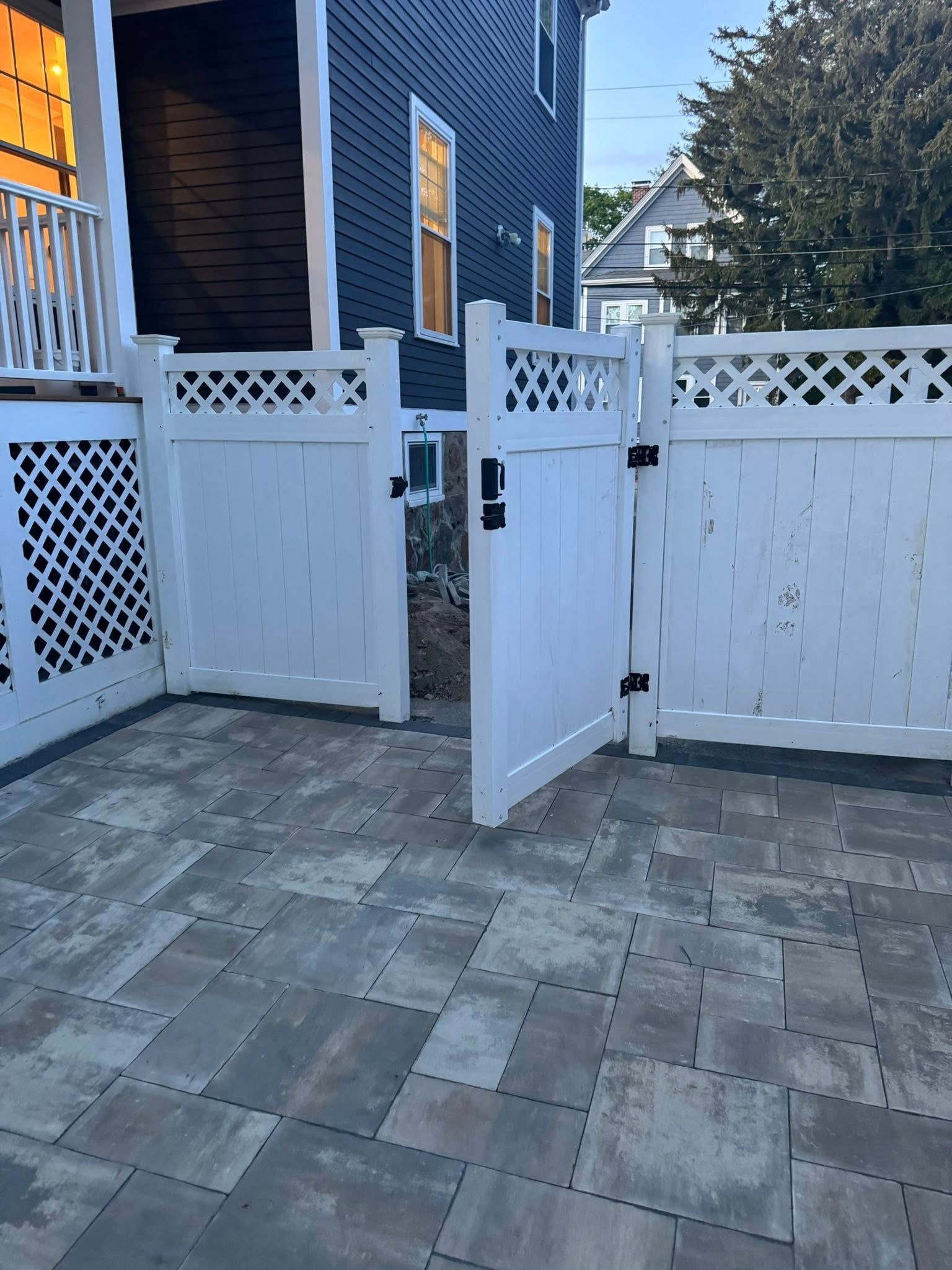 White vinyl fence with open gate on a gray paver patio, near a dark blue house.