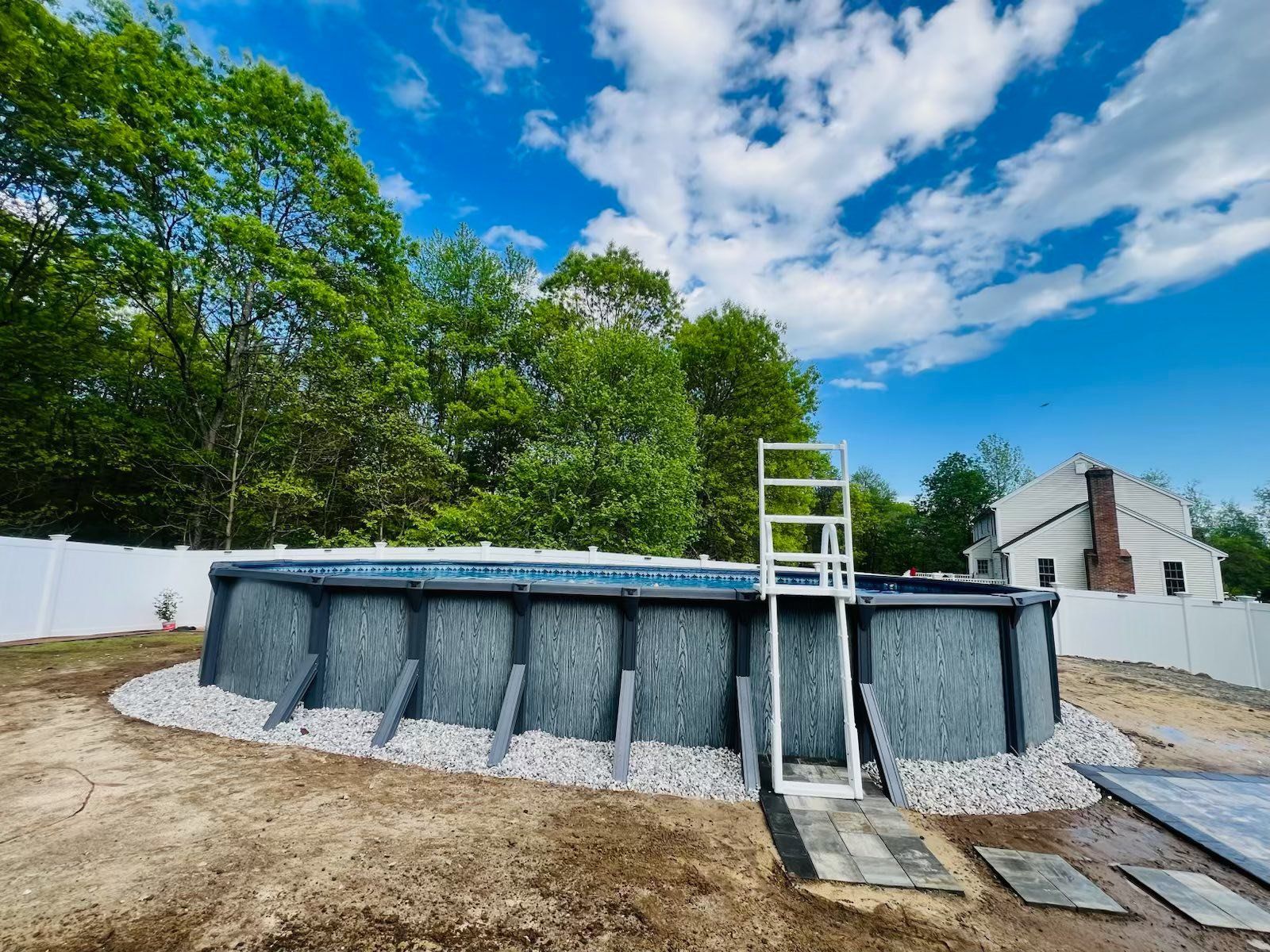 Above-ground pool with ladder, surrounded by gravel and a white fence, under a cloudy blue sky.
