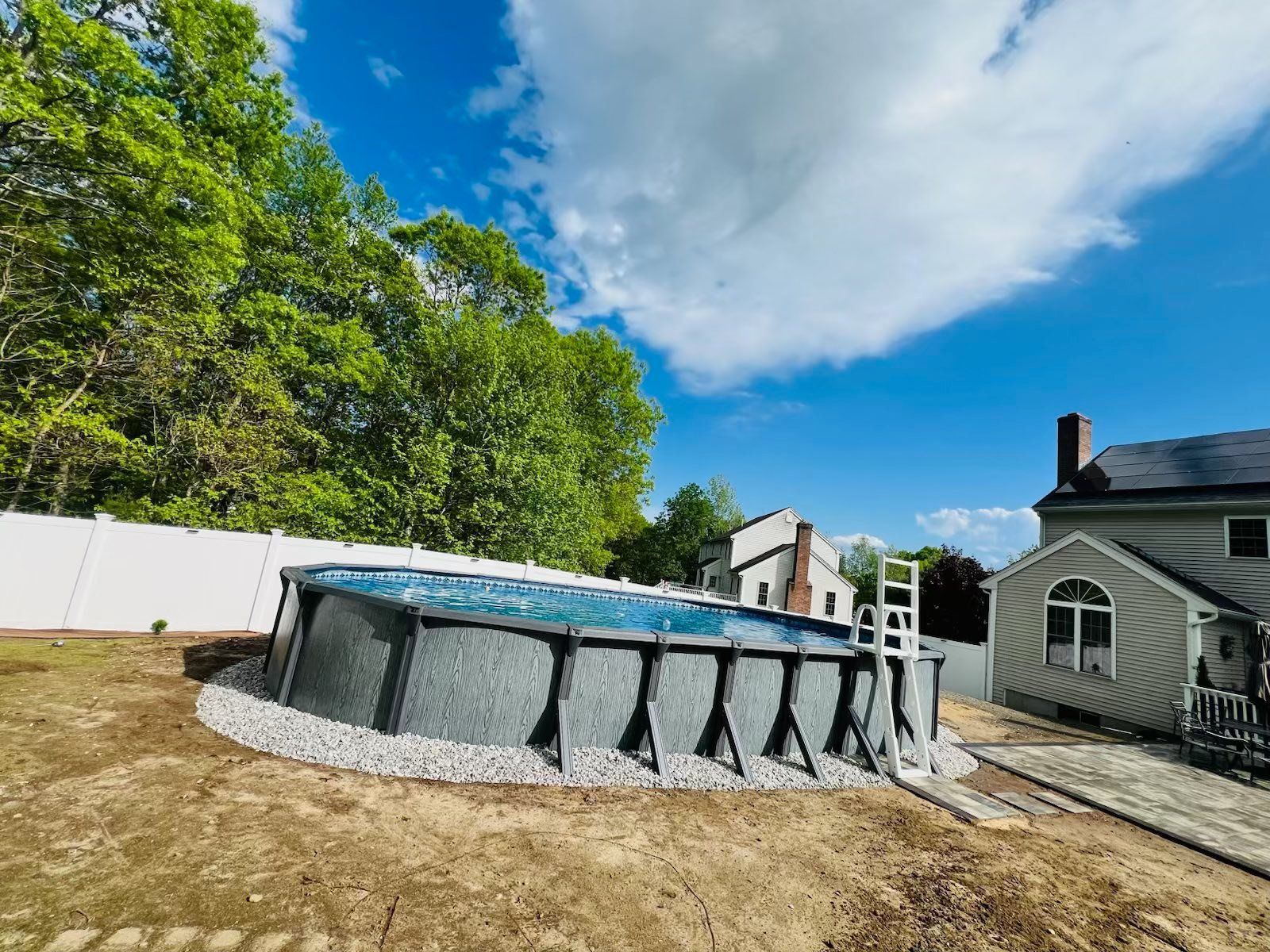 Above-ground pool in a backyard, surrounded by gravel, with trees and a house in the background on a sunny day.