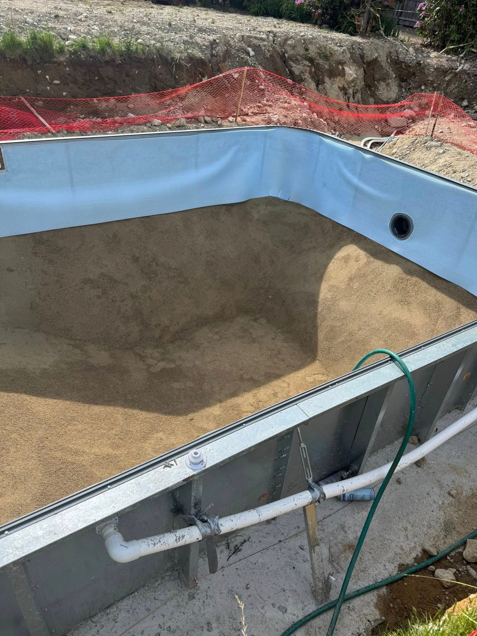 Pool under construction, steel walls, beige bottom, white plumbing, green hose, orange fencing.