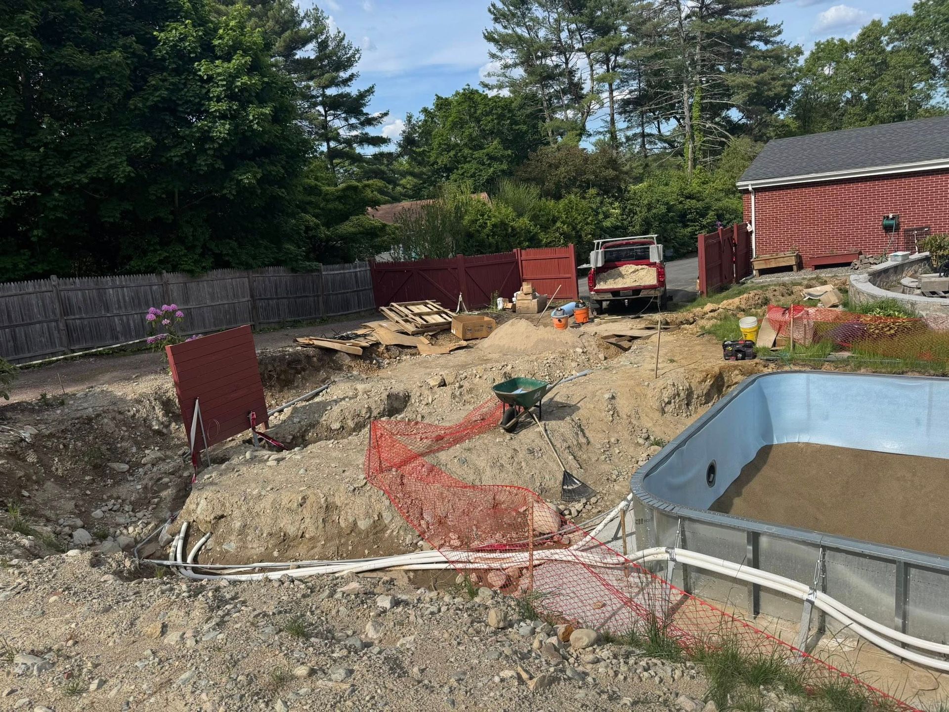 Pool being constructed; excavated ground with orange safety fencing, wheelbarrow, and truck.