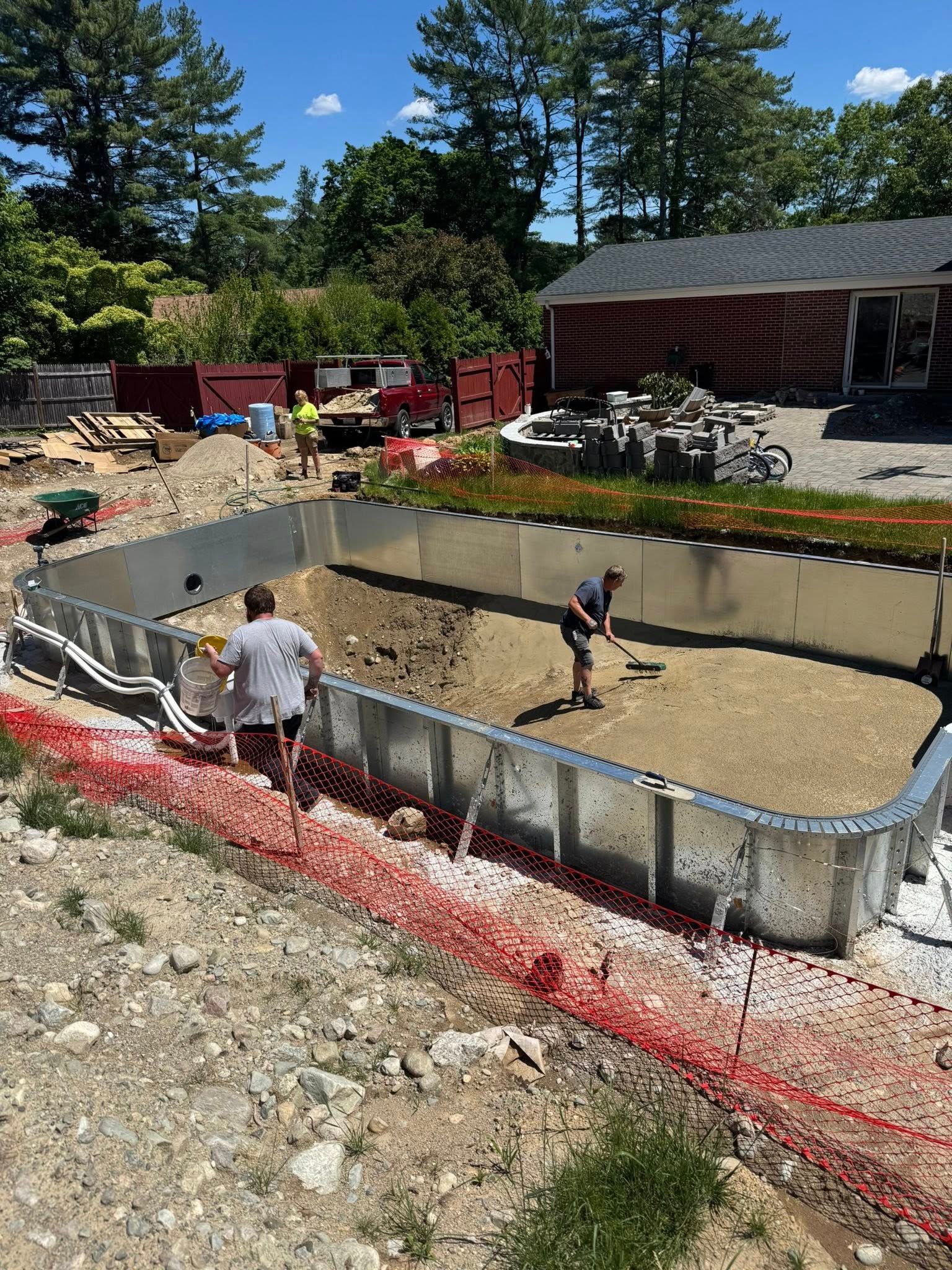 Pool construction site with workers. Metal frame, dirt, red safety fencing, sunny outdoors.