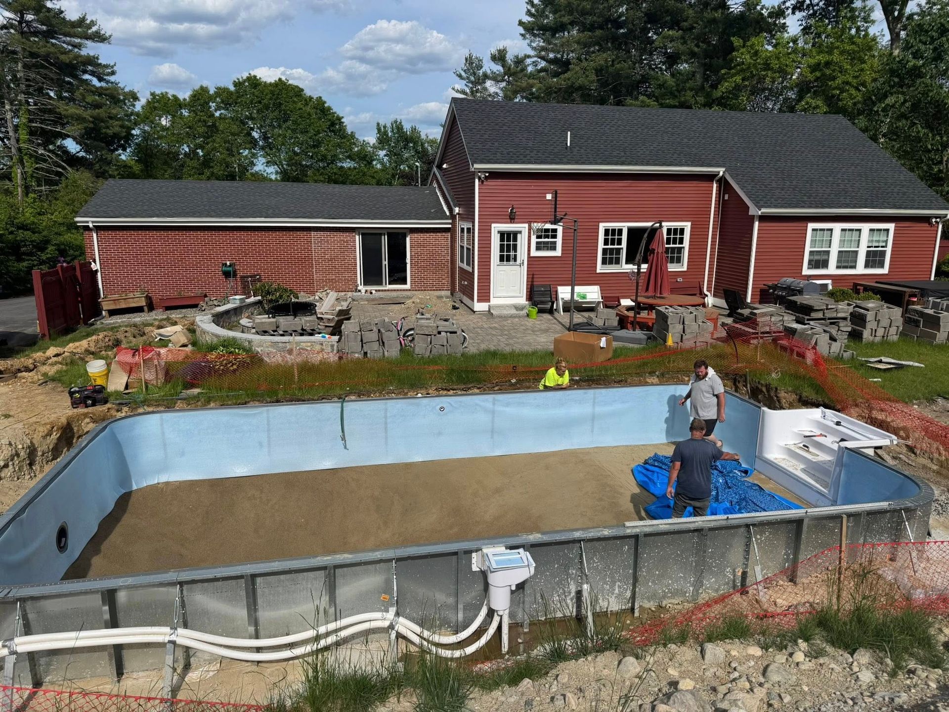 Pool construction: Workers in pool, house in background. Red house, blue pool liner, sand, and stone.