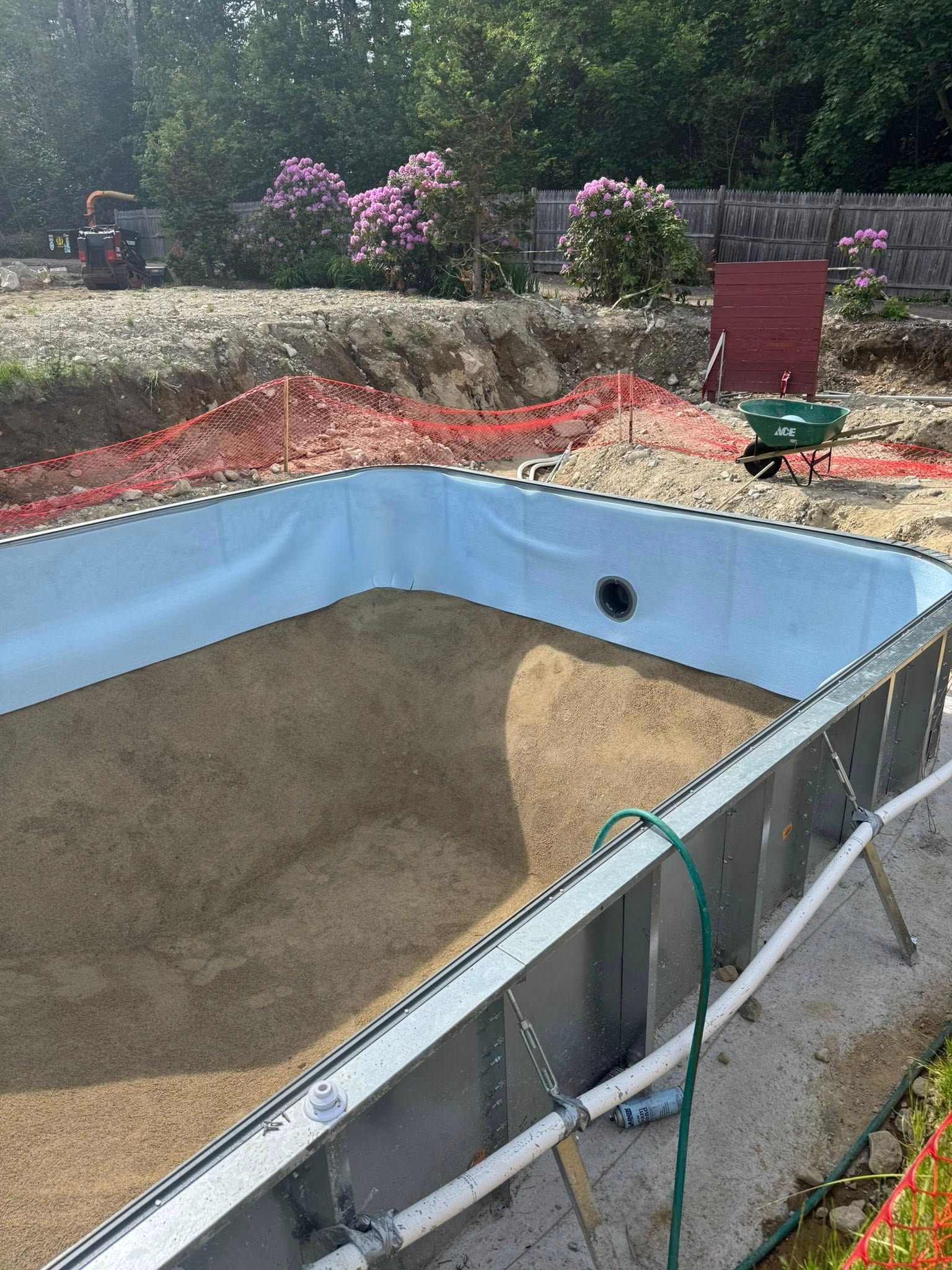 Pool under construction, steel frame, blue liner, filled with sand, surrounded by dirt and orange safety fencing.