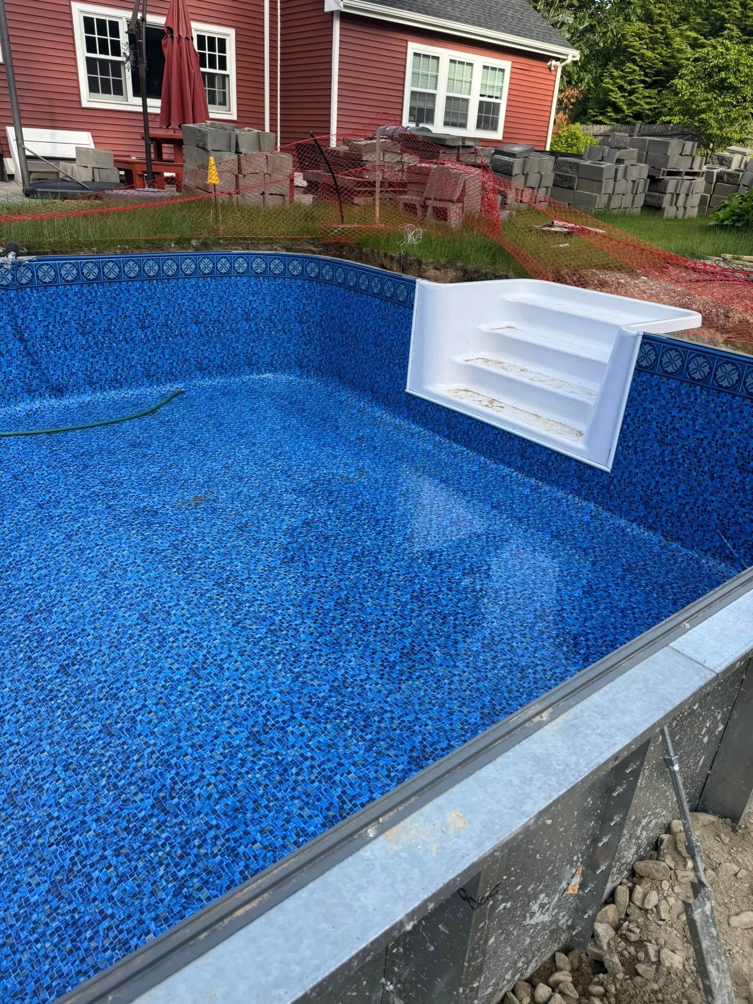 Blue-tiled swimming pool under construction, with white steps installed and a red house in the background.