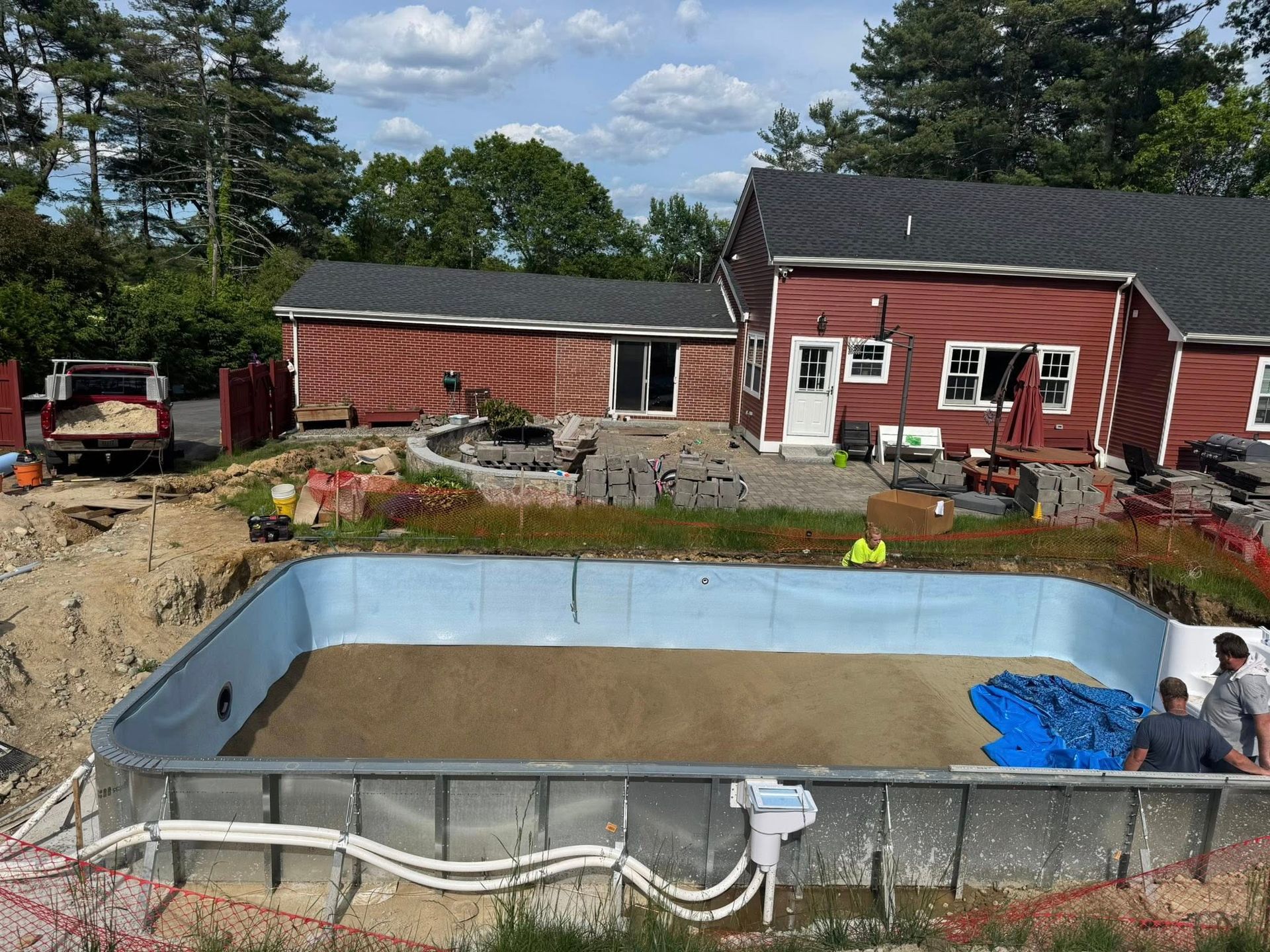Pool under construction with sand base, next to a red house and shed. Two workers, blue tarp.