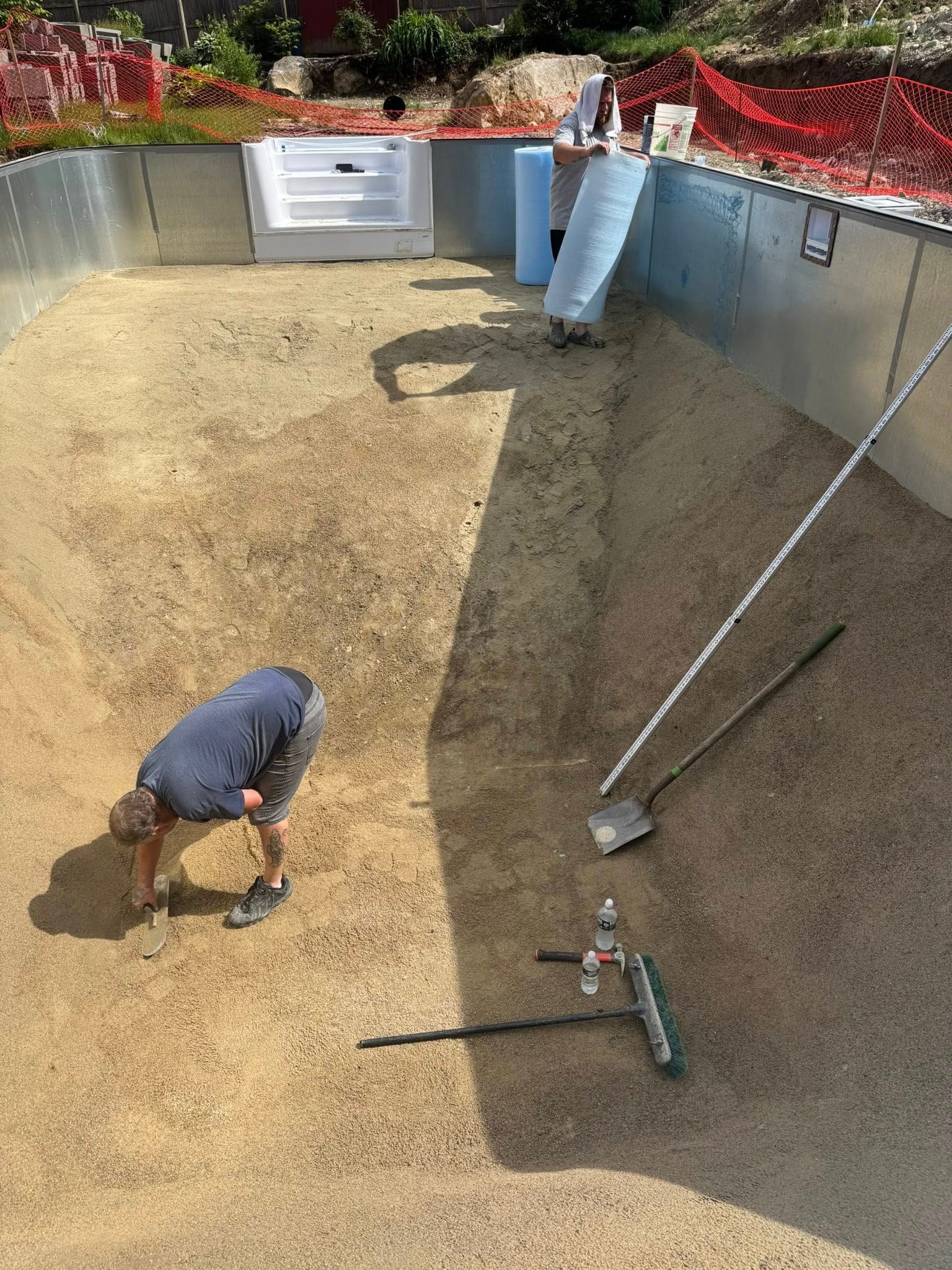 Man working inside a partially constructed pool, raking sand. Metal pool walls, white steps, tools present.
