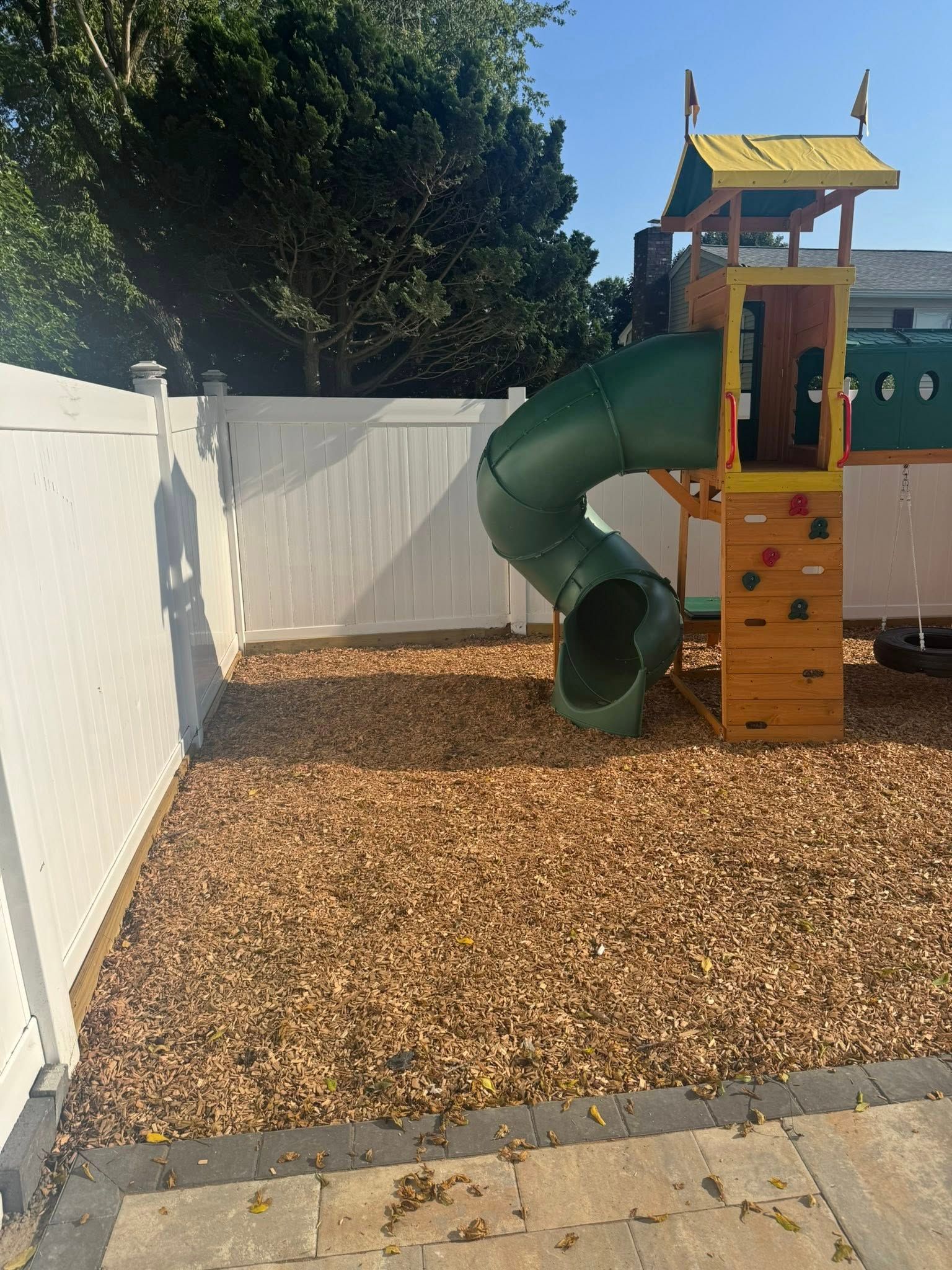 Playground with a green slide, climbing wall, and wood chip ground cover, next to a white fence.