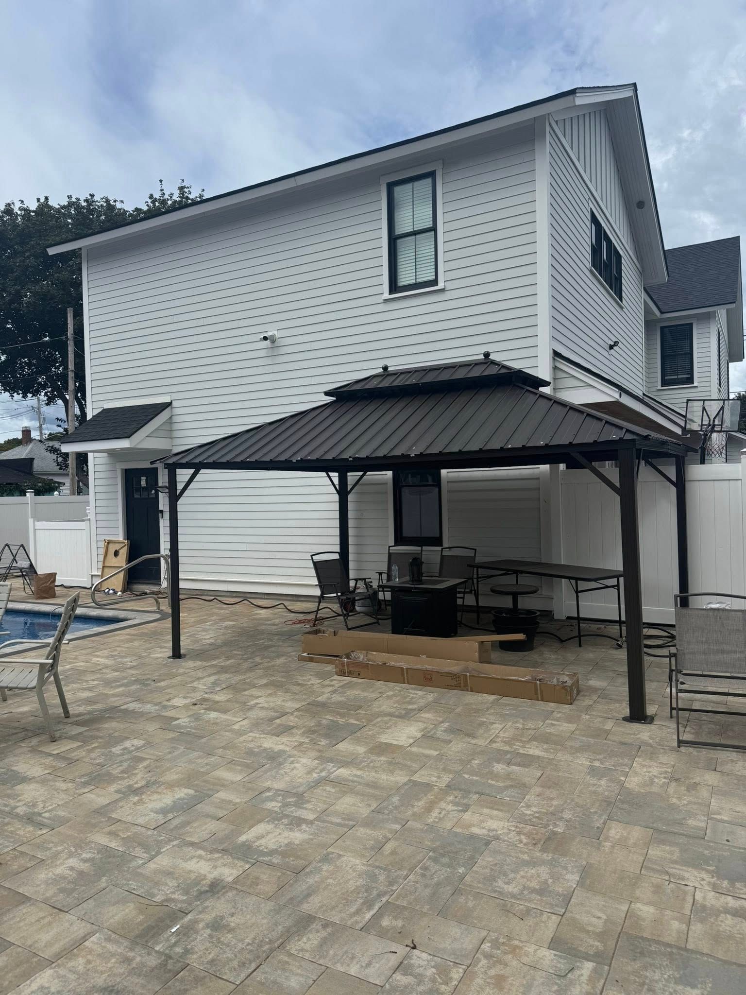 A backyard with a dark gazebo, pavers, a white house, and a swimming pool.
