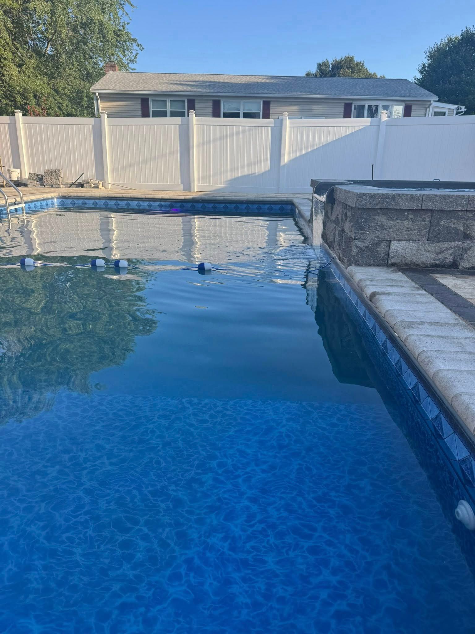 Pool with blue tiled surface, water, white fence, and house in background on a sunny day.